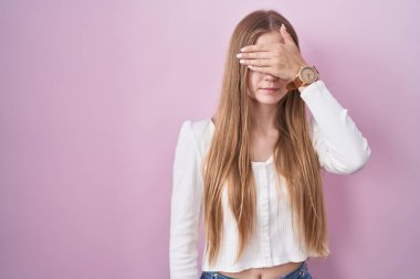 Young caucasian woman standing over pink background covering eyes with hand, looking serious and sad. sightless, hiding and rejection concept 