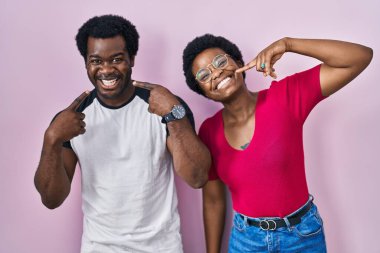 Genç Afro-Amerikalı çift pembe arka planda durup gülümseyip, parmaklarını ve ağzını gösteriyorlar. diş sağlığı kavramı. 