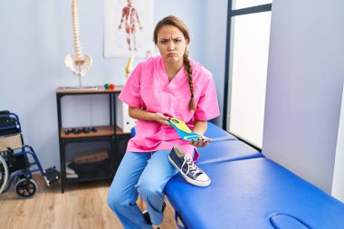 Fizyoterapi kliniğinde ayakkabı tabanı tutan genç İspanyol kadın şüpheci ve gergin, sorun yüzünden surat asıyor. negatif kişi. 