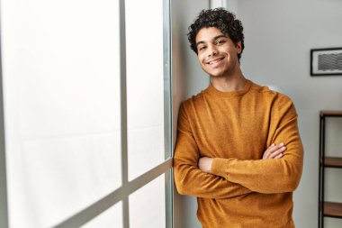 Genç İspanyol adam mutlu gülümsüyor. Kollarını kavuşturup evde duruyor..