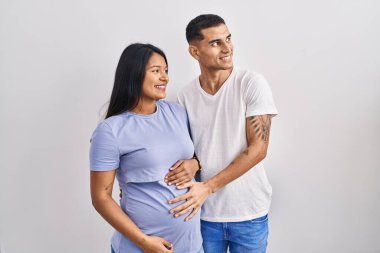 Latin kökenli genç bir çift arka planda bir bebek bekliyor. Yüzünde gülümseme, doğal bir ifade ile yan yana bakıyor. Kendinden emin gülen. 