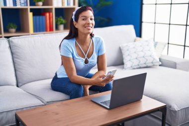 Young beautiful hispanic woman using smartphone and laptop at home