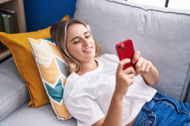 Evdeki kanepede uzanmış akıllı telefon kullanan genç sarışın kadın.