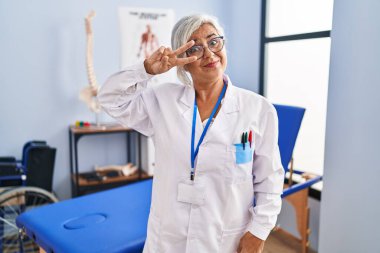 Middle age woman with grey hair working at pain recovery clinic doing peace symbol with fingers over face, smiling cheerful showing victory 