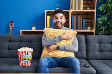 Hispanic man with beard eating popcorn watching a movie at home smiling and laughing hard out loud because funny crazy joke. 