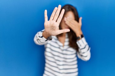 Young hispanic woman standing over blue isolated background covering eyes with hands and doing stop gesture with sad and fear expression. embarrassed and negative concept. 