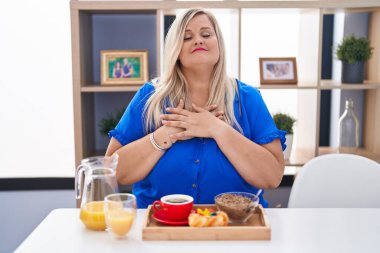Caucasian plus size woman eating breakfast at home smiling with hands on chest with closed eyes and grateful gesture on face. health concept. 