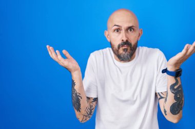 Mavi arka planda dikilen İspanyol dövmeli, elleri ve kolları havada, kafası karışmış bir adam. şüphe kavramı. 