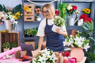 Young blonde woman florist using laptop holding plant at florist shop