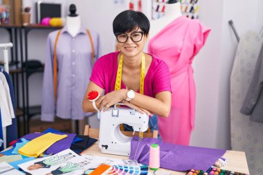 Middle age chinese woman tailor smiling confident leaning on sewing machine at atelier