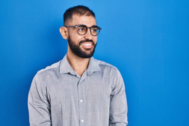 Middle east man with beard standing over blue background looking away to side with smile on face, natural expression. laughing confident. 