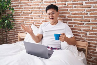 Young asian man drinking coffee sitting on the bed celebrating victory with happy smile and winner expression with raised hands 