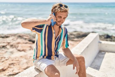 Young man tourist talking on the smartphone sitting on bench at seaside