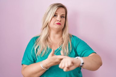 Caucasian plus size woman standing over pink background in hurry pointing to watch time, impatience, upset and angry for deadline delay 