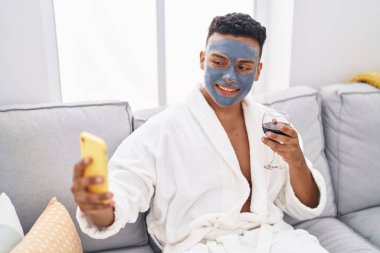 Young latin man sitting on sofa make selfie by smartphone drinking wine at home