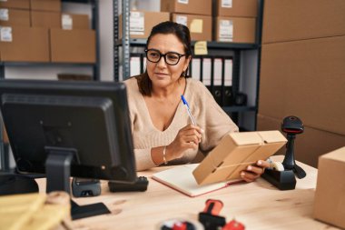 Middle age hispanic woman ecommerce business worker writing on notebook holding package at office