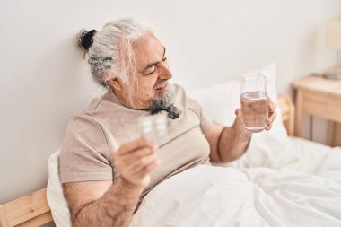 Middle age grey-haired man holding pills tablet and glass of water sitting on bed at bedroom