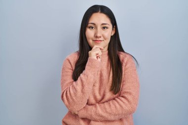 Young latin woman standing over blue background looking confident at the camera with smile with crossed arms and hand raised on chin. thinking positive. 