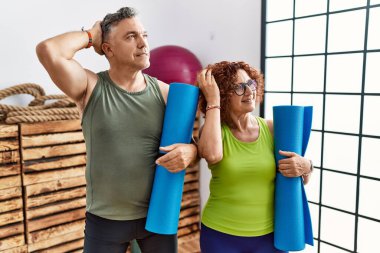 Orta yaşlı çift yoga minderi tutarken gülümsüyor. El hareketleriyle saçlarına dokunuyor, çekici ve modaya uygun poz veriyor. 