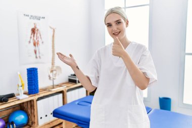 Ağrı kesici kliniğinde çalışan beyaz bir kadın avuç içini gösteriyor ve baş parmağıyla iyi bir hareket yapıyor, mutlu ve neşeli gülümsüyor. 