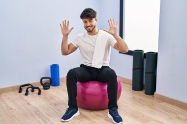 Sakallı İspanyol bir adam yoga odasında pilatese oturmuş parmaklarıyla on numarayı gösterirken kendinden emin ve mutlu bir şekilde gülümsüyor.. 