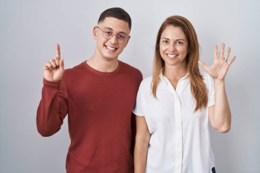 Anne ve oğlu izole edilmiş arka planda yan yana duruyorlar. Altıncı parmaklarıyla işaret ediyorlar ve gülümsüyorlar. Kendinden emin ve mutlular.. 