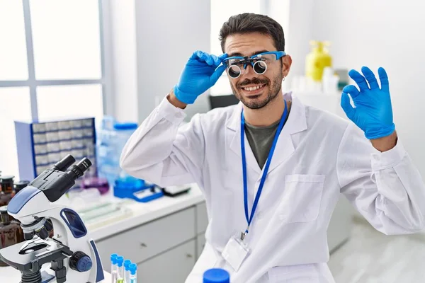 若いヒスパニック系の男は 指でOkサインを行う拡大鏡を身に着けている科学研究所で働いています 笑顔優しいジェスチャー優れたシンボル — ストック写真