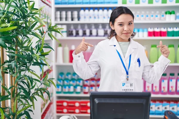 Hispanic young woman working at pharmacy drugstore looking confident with smile on face, pointing oneself with fingers proud and happy.