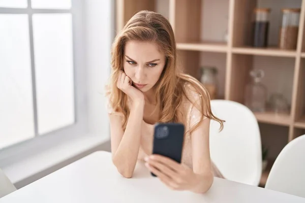 年轻的金发女人在家里用带有严肃表情的智能手机 — 图库照片