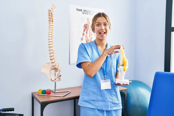 Młoda Blondynka Ortopedyczna Kobieta Trzyma Anatomiczny Model Stawu Kolanowego Świętując — Zdjęcie stockowe
