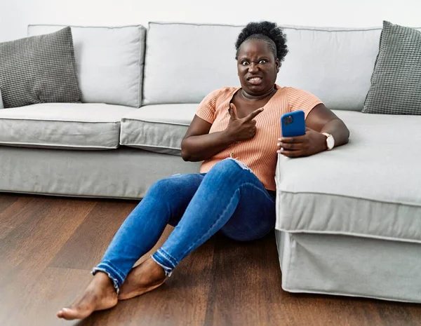 年轻的非洲女人用智能手机坐在家里的地板上 用食指 关心和惊讶的表情将焦虑和紧张抛在一边 — 图库照片