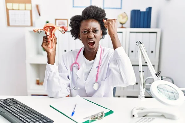 African doctor woman holding anatomical model of female genital organ at medical clinic annoyed and frustrated shouting with anger, yelling crazy with anger and hand raised