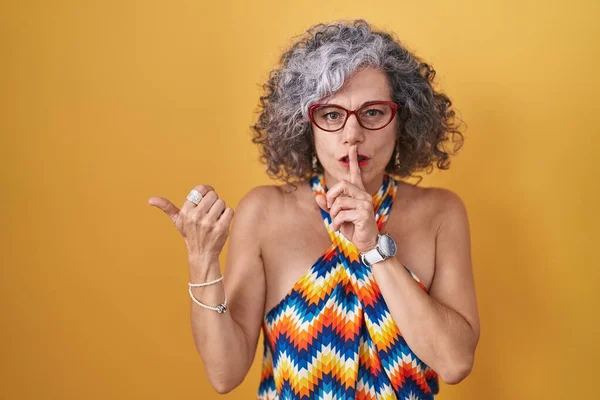 Middle age woman with grey hair standing over yellow background asking to be quiet with finger on lips pointing with hand to the side. silence and secret concept.
