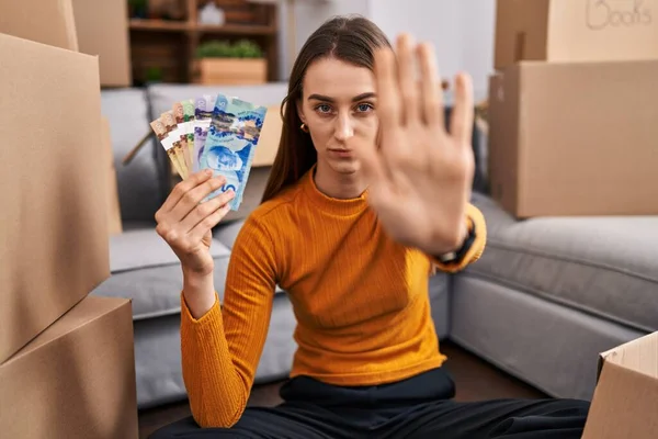 Young caucasian woman sitting on the floor at new home holding canadian money with open hand doing stop sign with serious and confident expression, defense gesture