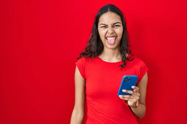 年轻的巴西女人在红色背景下用智能手机伸出舌头 带着滑稽的表情很高兴 情感概念 — 图库照片