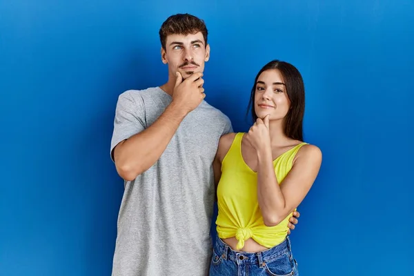Young hispanic couple standing together over blue background looking confident at the camera smiling with crossed arms and hand raised on chin. thinking positive.