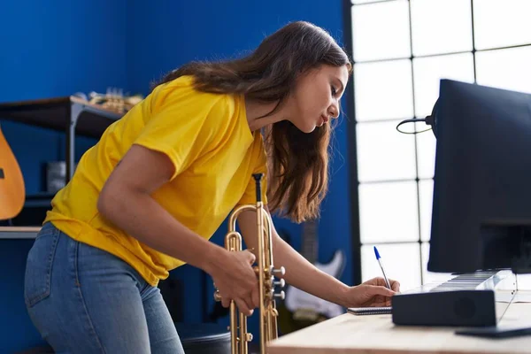 Young woman musician holding trumpet writing on notebook at music studio