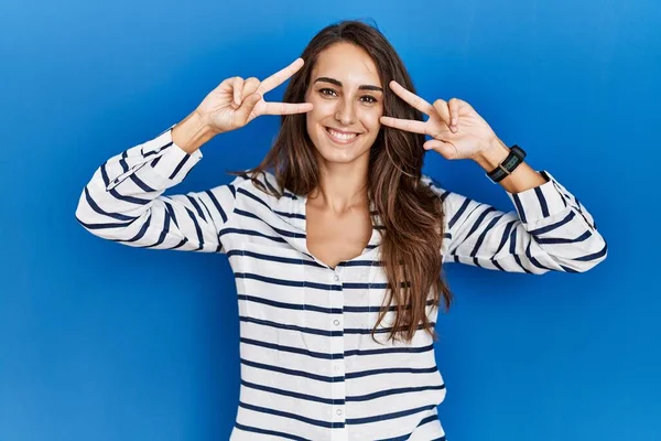 Young hispanic woman standing over blue isolated background doing peace symbol with fingers over face, smiling cheerful showing victory