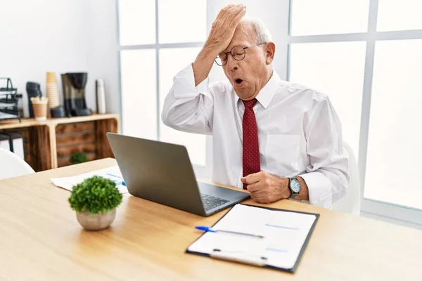 Senior man working at the office using computer laptop surprised with hand on head for mistake, remember error. forgot, bad memory concept.