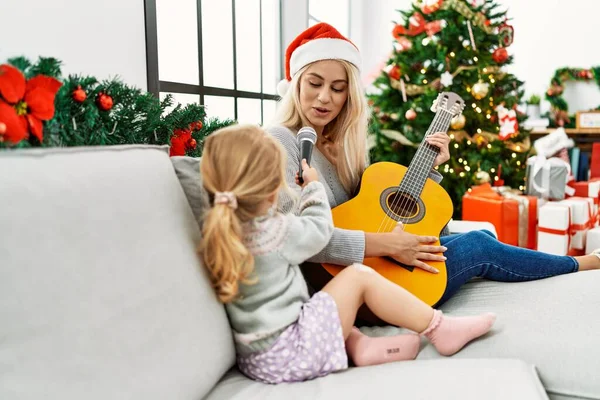 Mother and daughter playing guitar and singing sitting by christmas tree at home