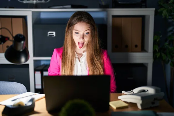 年轻的高加索女人晚上在办公室里工作 带着滑稽的表情伸出舌头 高兴极了 情感概念 — 图库照片