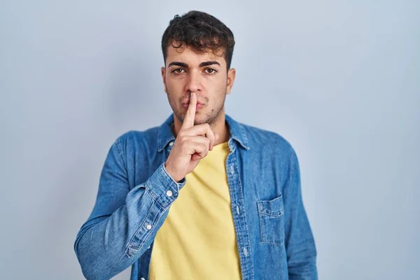 青い背景の上に立っている若いヒスパニック系の男が唇に指で静かにするように求めている 沈黙と秘密の概念 — ストック写真