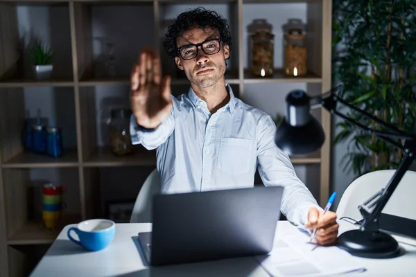 Hispanic man working at the office at night doing stop sing with palm of the hand. warning expression with negative and serious gesture on the face.