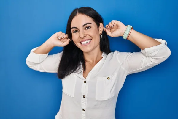 若いヒスパニック系の女性の青い背景に指で耳を引く笑みを浮かべて立って 面白いジェスチャー オーディション問題 — ストック写真