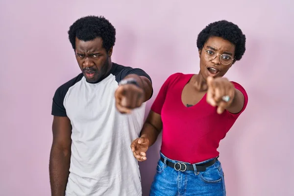 Young african american couple standing over pink background pointing displeased and frustrated to the camera, angry and furious with you