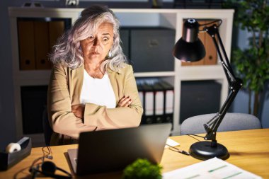 Gri saçlı orta yaşlı bir kadın gece geç saatlerde bilgisayar laptopu kullanıyor şüpheci ve gergin, yüzünde çapraz kollu tasvip etmeyen bir ifade var. negatif kişi. 