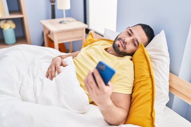 Young latin man using smartphone lying on bed at bedroom