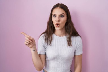 Young hispanic girl standing over pink background surprised pointing with finger to the side, open mouth amazed expression. 