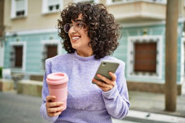 Orta Doğulu genç bir kadın sokakta akıllı telefon kullanarak gülümsüyor.