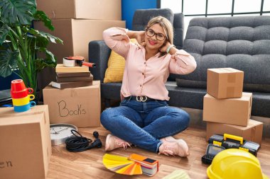 Young hispanic woman moving to a new home sitting on the floor relaxing and stretching, arms and hands behind head and neck smiling happy 